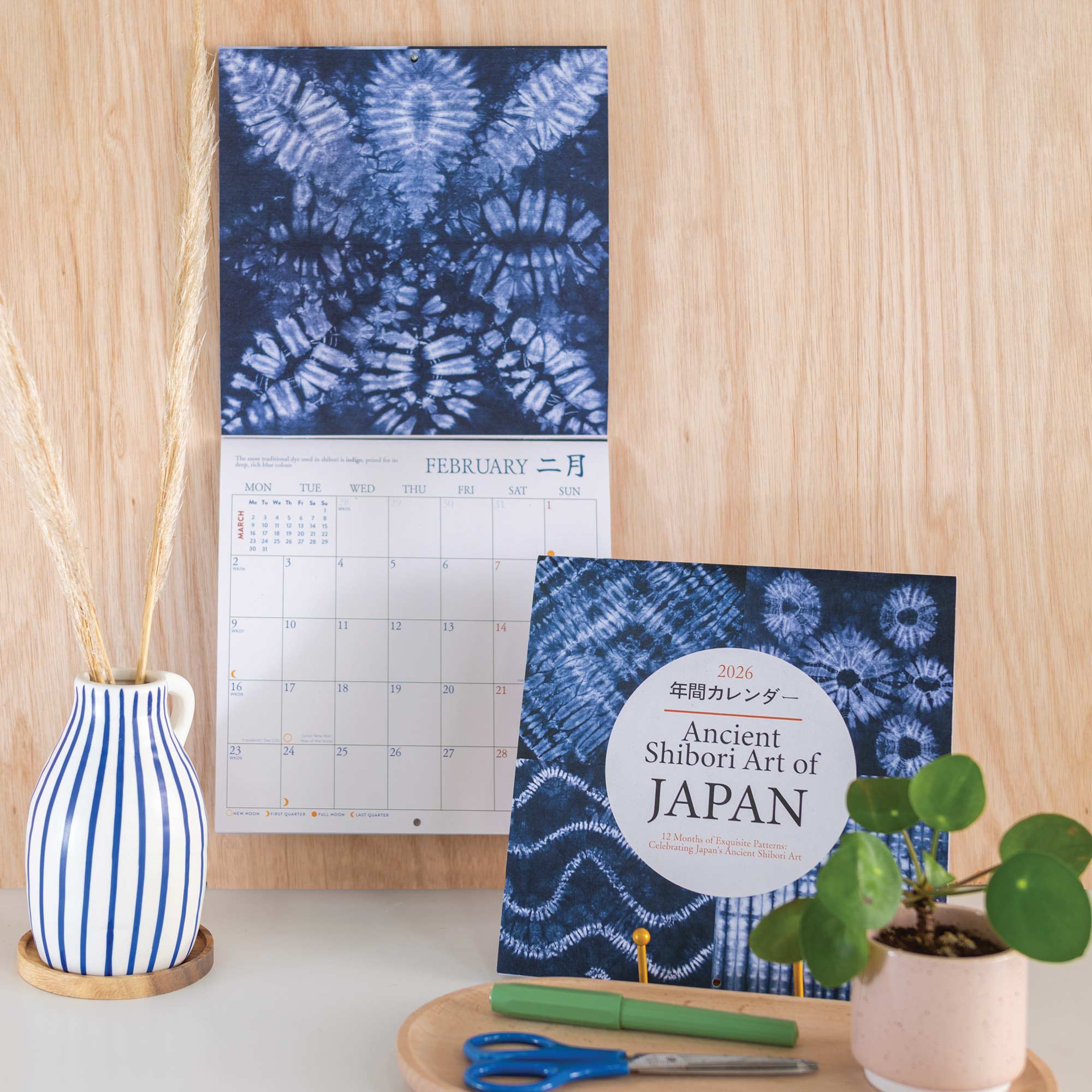 Calendar with blue tie-dye design and a book titled 'Ancient Shibori Art of Japan' on a wooden surface.