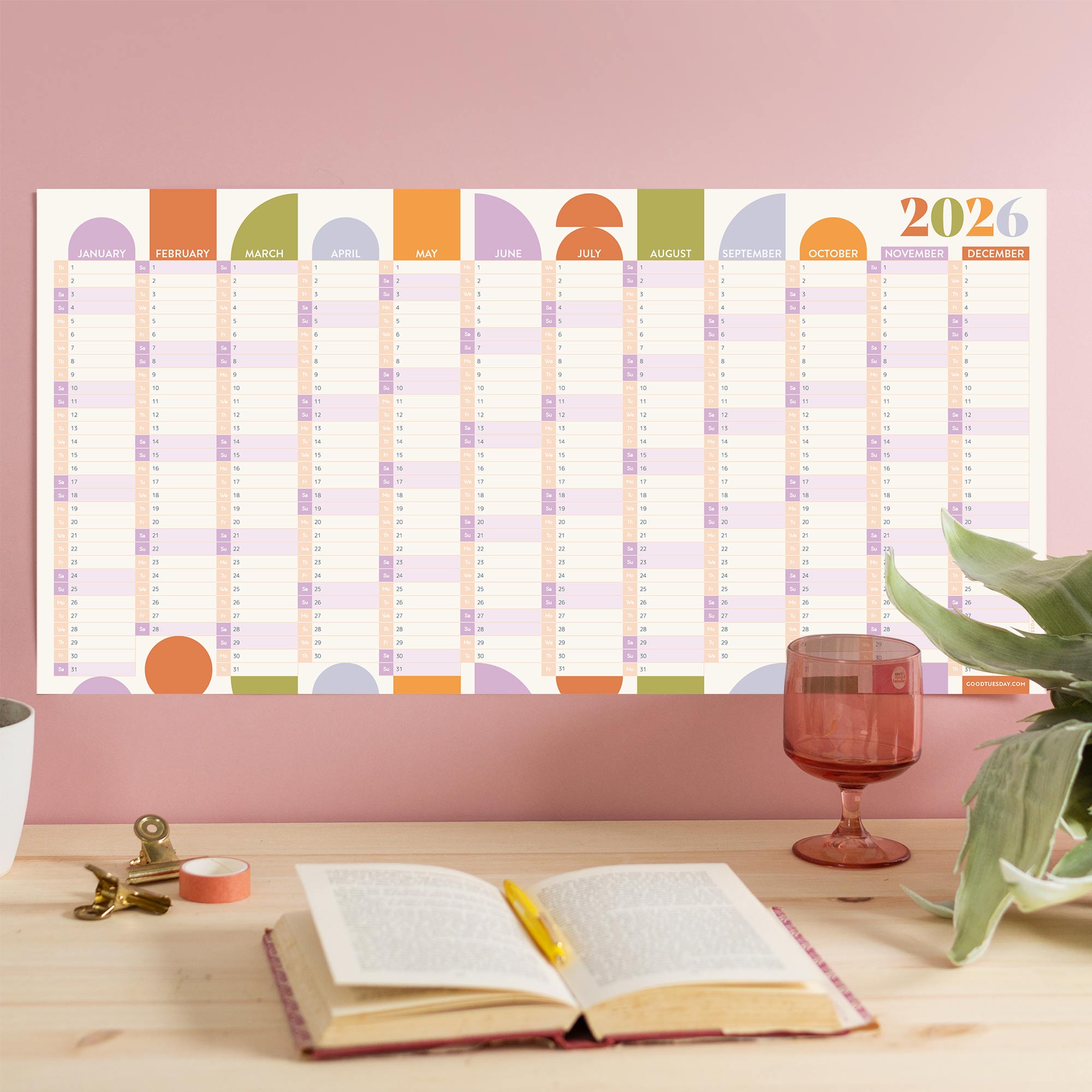 A full view of a "Geometric Citrus Splash" 2026 wall planner, laid out flat on a light wooden desk with a plant, a book, and a glass visible. The planner features a colorful, minimalist design with all months of the year displayed horizontally.