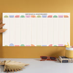 A pastel-themed "Birthdays & Anniversaries" wall planner displayed above a desk. The planner shows all twelve months with ample space to write.