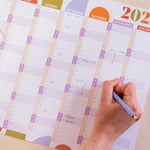 A close-up of a hand writing on a 2026 wall planner. Weekends are highlighted in a different colour.