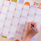 A close-up of a hand writing on a 2026 wall planner. Weekends are highlighted in a different colour.