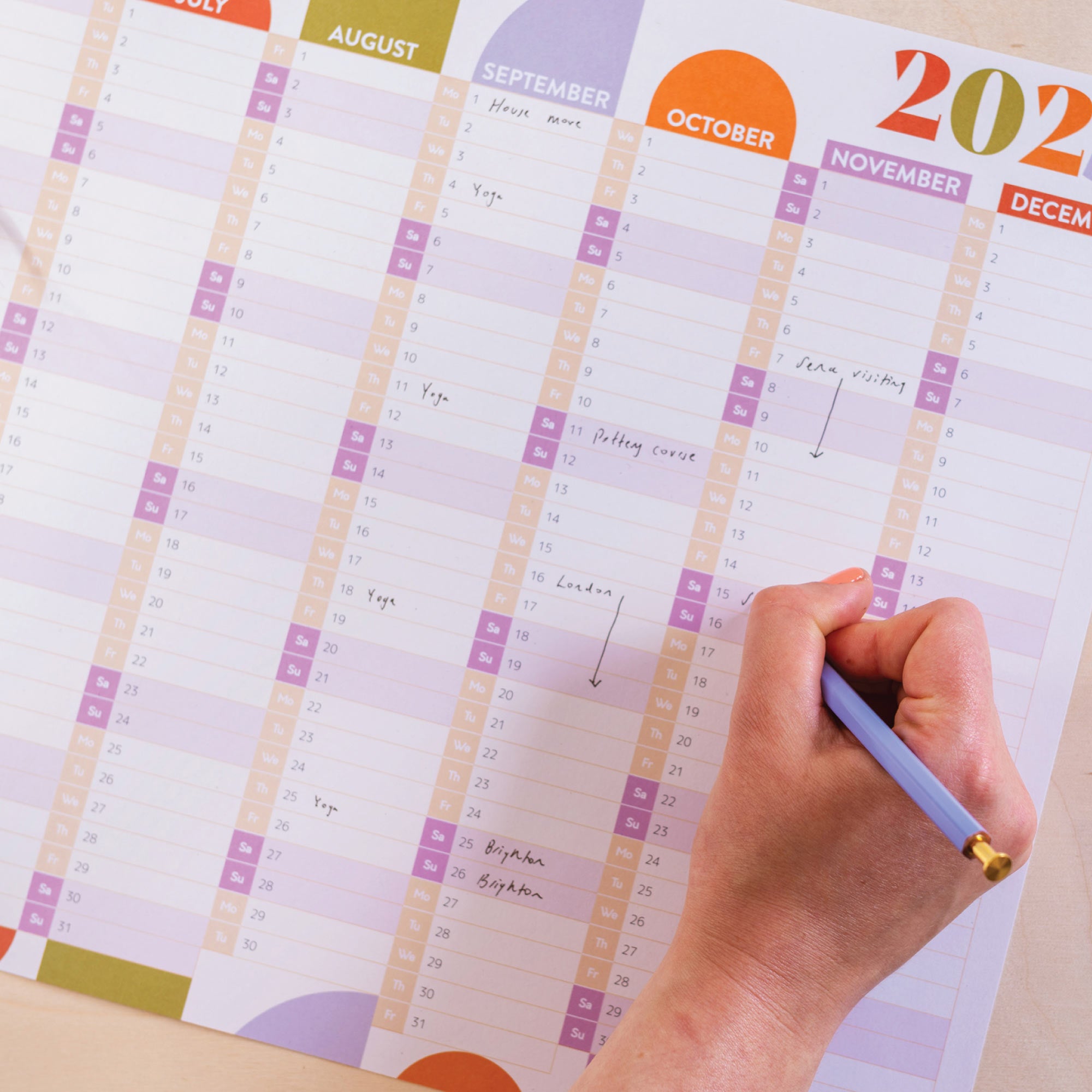 A close-up of a hand writing on a 2026 wall planner. Weekends are highlighted in a different colour.