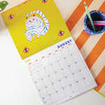 2026 bright and colourful Calendar with a Cat artwork by the artist Ashley Percival. Showing the Page open on the month of August with a bright yellow illustration of a cat.