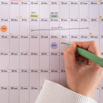 Close up of hand with a green pen writing on Good Tuesday's 2026 Black and White wall calendar with events marked. Good Tuesday's 2026 black and white landscape wall planner. 100% Recycled paper. 