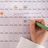 Close up of hand with a green pen writing on Good Tuesday's 2026 Black and White wall calendar with events marked. Good Tuesday's 2026 black and white landscape wall planner. 100% Recycled paper. 