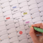 Hand holding a green pen over Good Tuesday's Black and White 2026 calendar with events marked. 100% recycled Paper, Made in the UK.