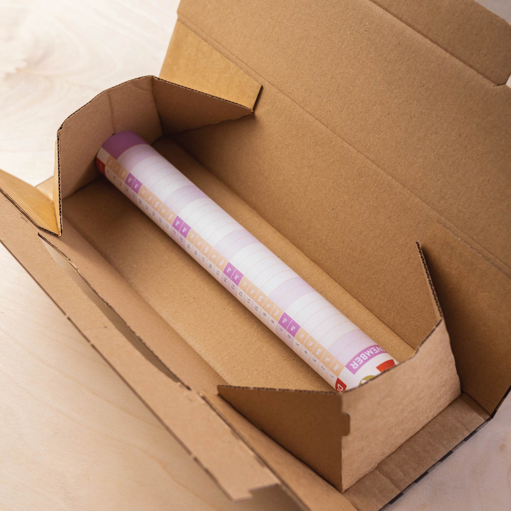 A rolled-up 2026 wall planner, featuring a purple and orange design, shown rolled inside a brown cardboard box, ready for shipping.