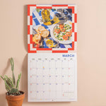 Calendar with a Mediterranean theme on a beige wall, featuring images of food and drinks.