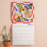 Calendar with a Mediterranean theme on a beige wall, featuring images of food and drinks.