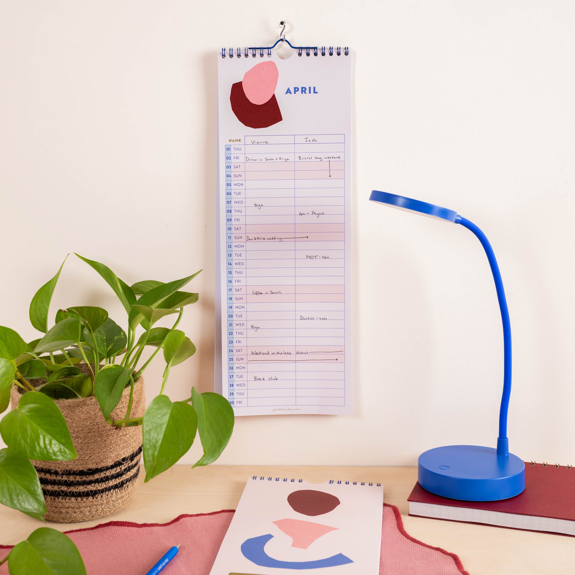 A spiral-bound April couples calendar with abstract color blocks hangs on a wall above a desk.