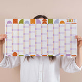 Person holding a compact pastel 2027 wall planner with colourful squares on a beige background