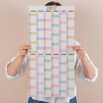 Person holding a compact pastel 2027 wall planner with colourful squares on a beige background