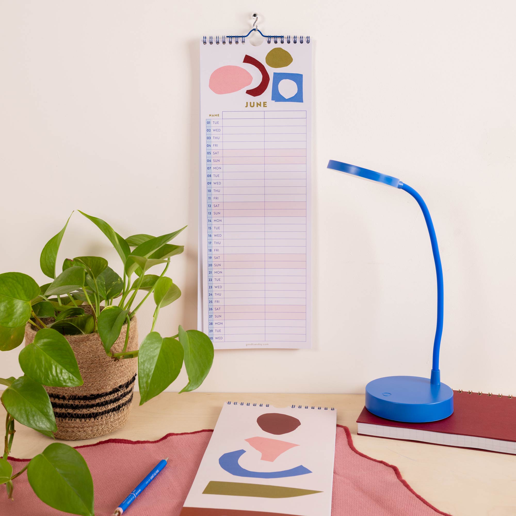 A spiral-bound June calendar page with abstract pastel shapes hangs on a wall above a desk.