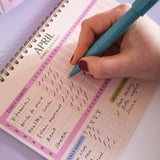 Hand with a teal pen writing in a pink habit tracker journal.