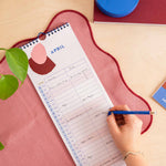 A spiral-bound monthly planner pad labeled April is laid on a pink textured mat on a light wood desk, with a hand writing in it with a blue pencil.