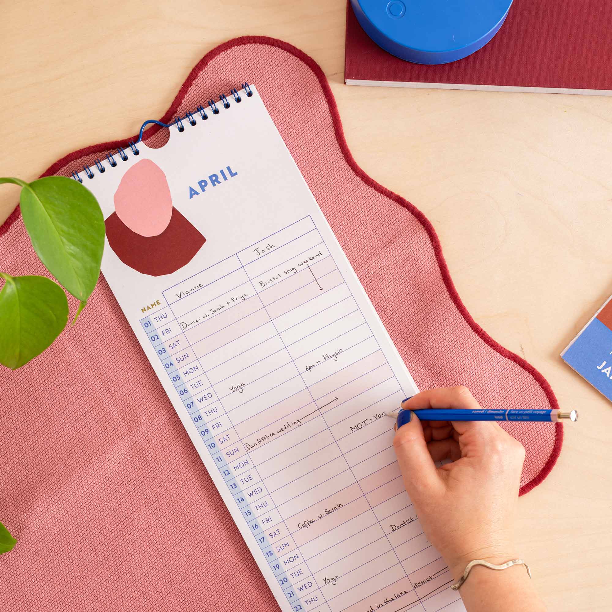 A spiral-bound monthly planner pad labeled April is laid on a pink textured mat on a light wood desk, with a hand writing in it with a blue pencil.