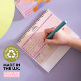 Hand writing in a planner with a pen on a purple surface, featuring recycled paper and plastic-free packaging text.