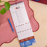 A spiral-bound planner or calendar pad titled 'Trying to Co-Ordinate My Busy Schedules!' lies on a pink fabric surface over a light wood tabletop.