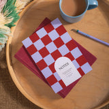 Plum and lilac checkered notebook with 'NOTES' on a wooden tray with a cup of coffee and pen.