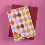 Two notebooks, one with a colorful diamond pattern cover and the other plain red, on a pink background.