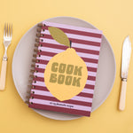 Lemon Zest Recipe Journal with lemon graphic on top of Plum and Lilac stripes on the cover design, placed on a plate with utensils on a yellow background