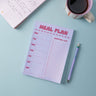 Meal planner pad and shopping list notepad with a pen and coffee cup on a light blue background