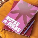 Pink notebook with 'NOTES & IDEAS' text on a yellow surface