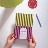 Pisatchio Striped notebook with 'NOTES' label held by hands on a white surface with a smiley face coaster and pen.