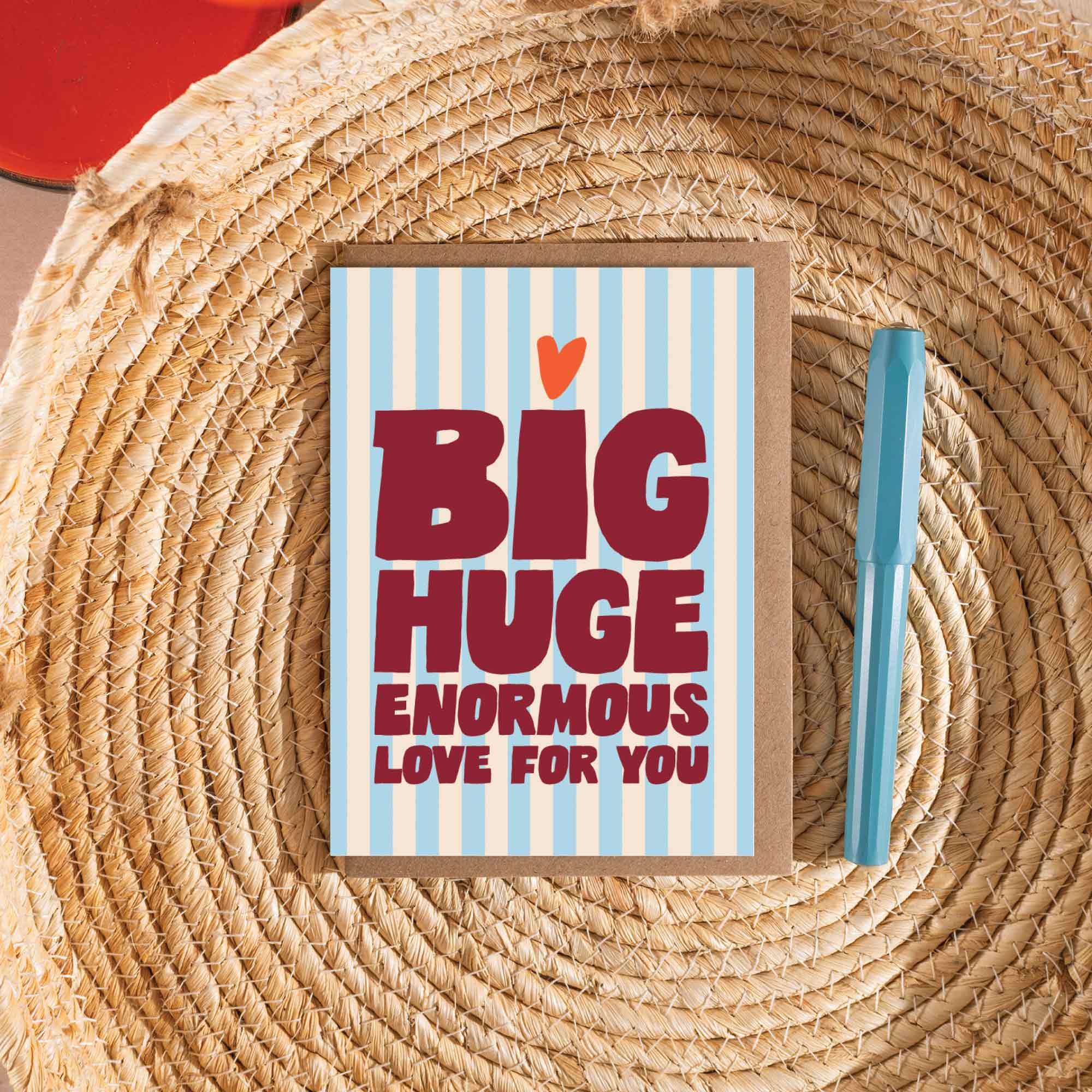 Card with 'BIG HUGE ENORMOUS LOVE FOR YOU' text on a glacial blue and cream striped background, placed on a woven surface with a pen.