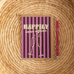 Greeting card with 'Happily ever after' text and champagne flutes on a striped background, accompanied by a pen on a woven surface.