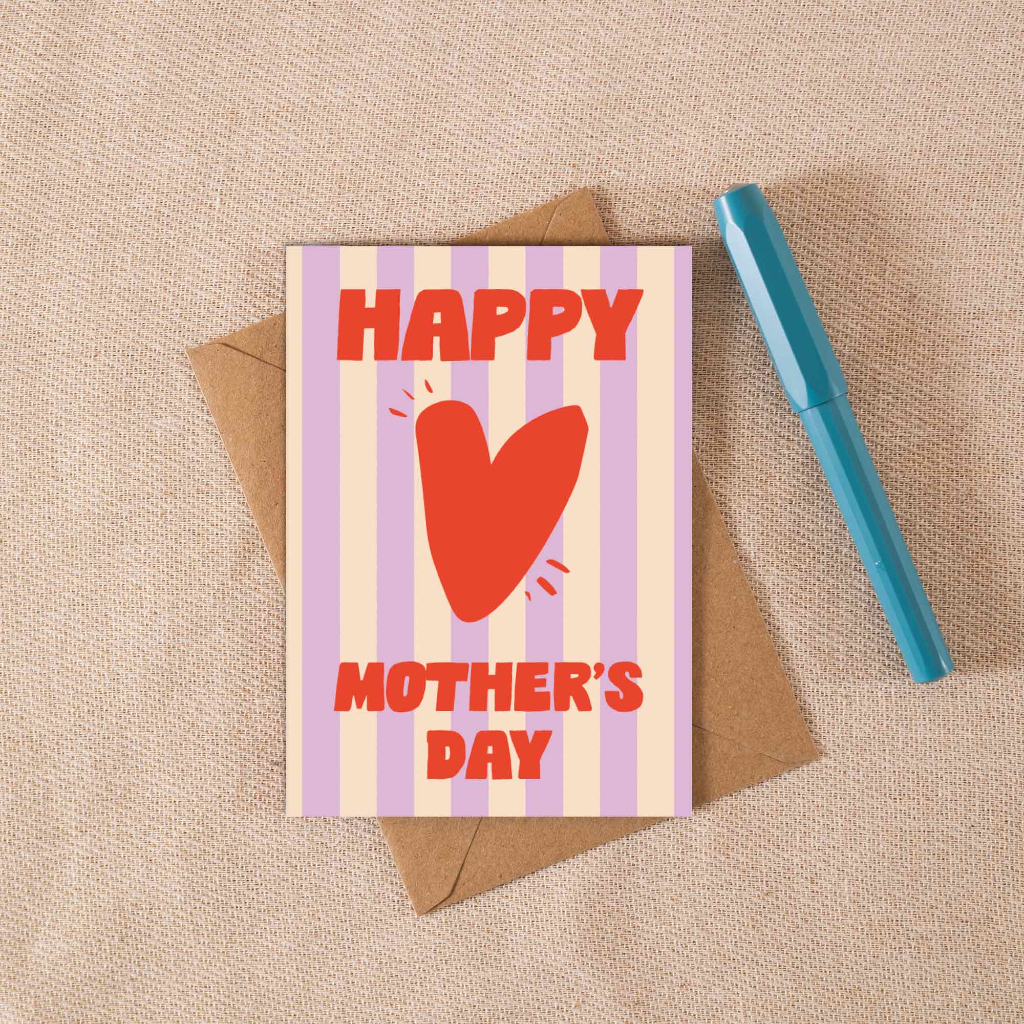 Mother's Day card with red heart and text on striped background, placed on a textured surface with a pen.