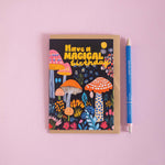 Greeting card with colorful mushrooms and text 'Have a Magical Birthday' on a pink background, next to a pen.
