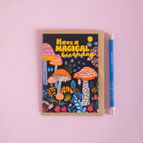Greeting card with colorful mushrooms and text 'Have a Magical Birthday' on a pink background, next to a pen.