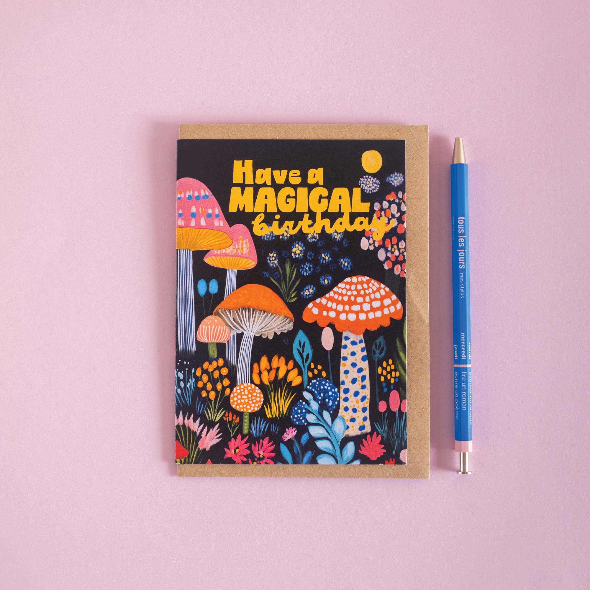 Greeting card with colorful mushrooms and text 'Have a Magical Birthday' on a pink background, next to a pen.