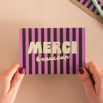 Purple and pink striped card with 'MERCI beaucoup' text held by hands against a beige background