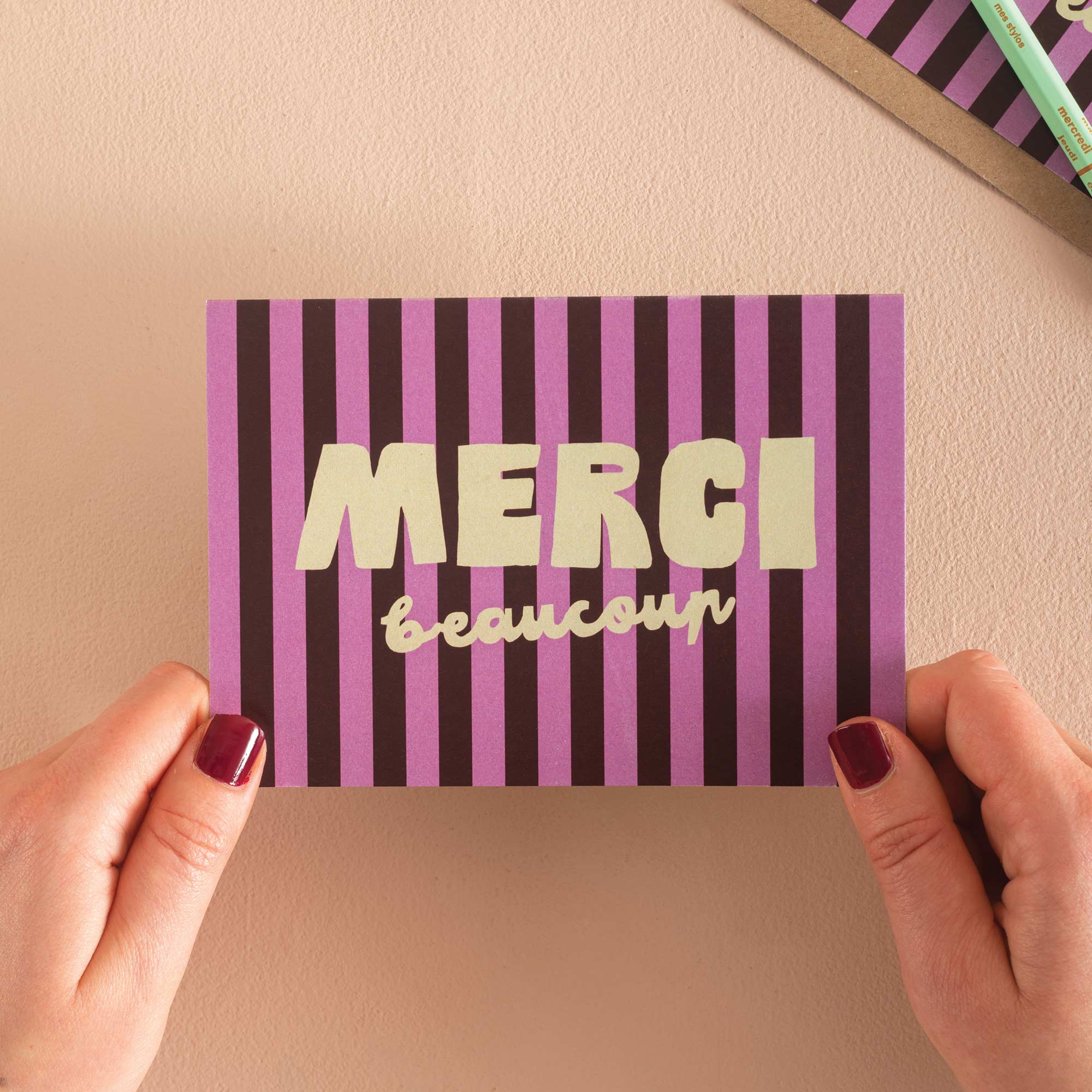 Purple and pink striped card with 'MERCI beaucoup' text held by hands against a beige background