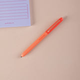 Orange ball point pen on a beige surface with a purple notepad in the corner.