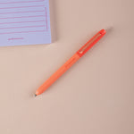 Orange ball point pen on a beige surface with a purple notepad in the corner.