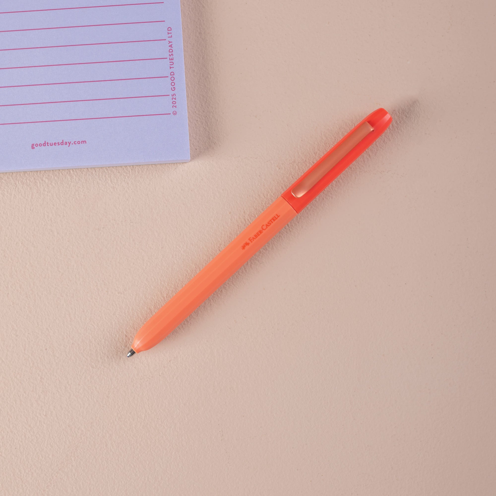 Orange ball point pen on a beige surface with a purple notepad in the corner.