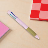 Multicolored pink pen on a beige surface with a red checkered notebook in the background.