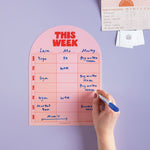 Pink weekly planner with a hand writing on it against a purple wall.